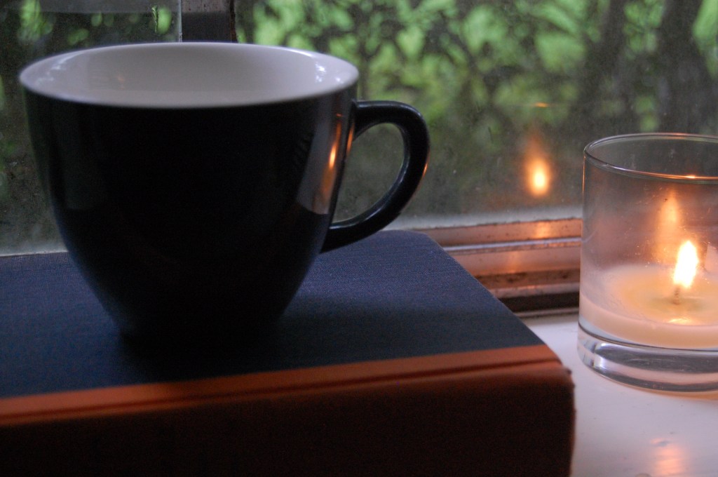 A gray mug on a book with an orange-colored spine; a mostly empty candle burns to the right, reflecting against the mug and a window pane.