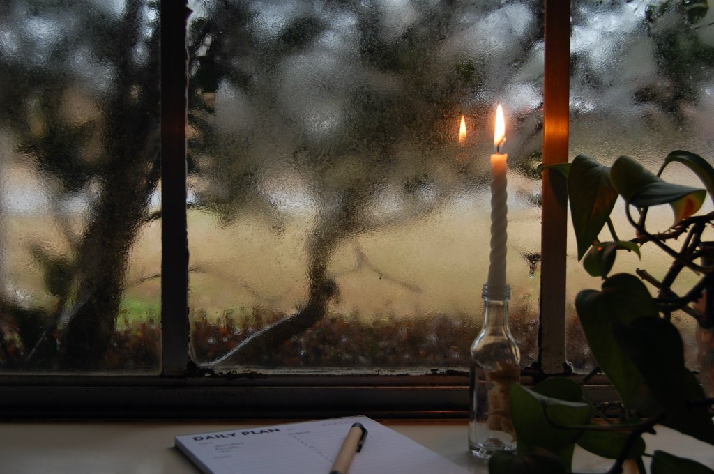 A planner and pen next to a thin, twisted burning candle in a tiny glass bottle, and a green plant; in the background, a window to a brown lawn and bushes blurred by rain