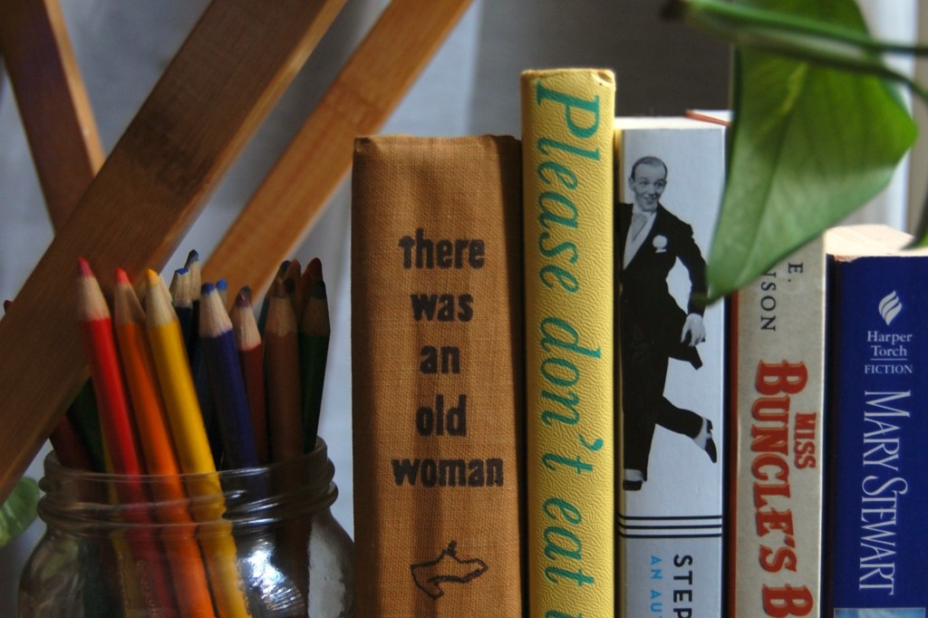 a jar of colored pencils next to five books (color of spines are: brown, yellow, black/white, tan/red, purple). A green leaf at top right, slanted bars of natural wood at top left.